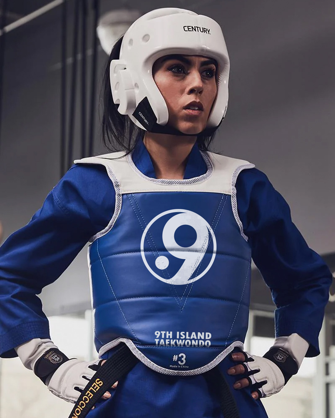 Woman wearing white Century headgear and blue 9th Island Taekwondo chest guard with black belt and white hand gloves.